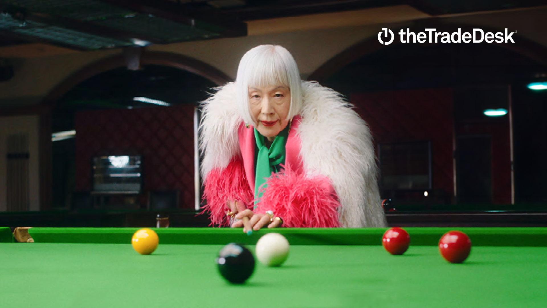 A woman wearing a pink and white fur jacket plays pool